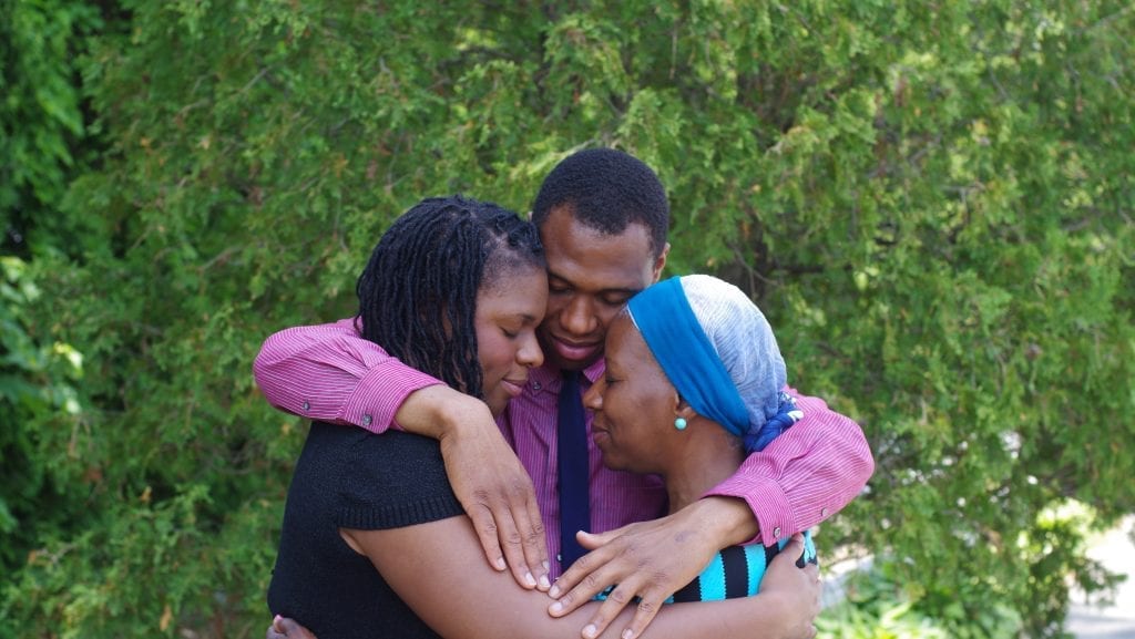 Son embraces mom and sister with a green shrub in the background.