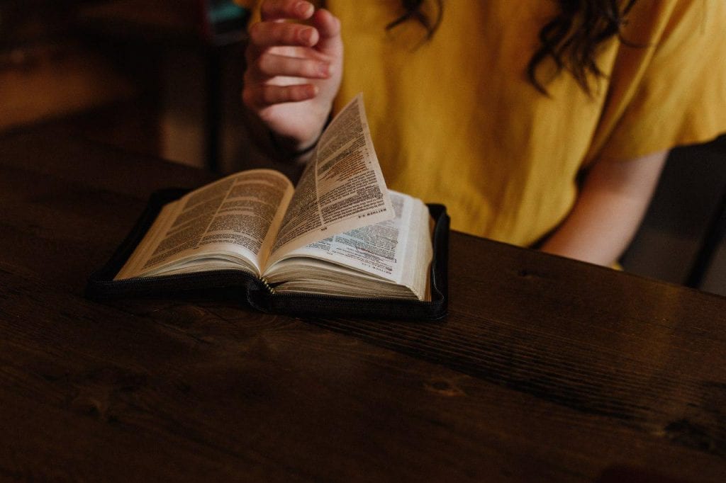 A bible on top of a table with it open and a woman flipping through the pages.
