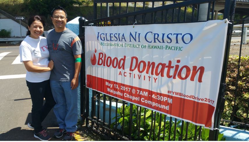 A couple smiling together outdoors in front of an iron gate and banner that reads Blood Donation Activity.