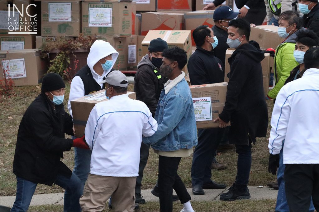Iglesia Ni Cristo (INC or Church of Christ) members carry the donation boxes one by one outdoors with masks on, complying with the COVID-19 government restrictions, with the stacked donation boxes in the background.