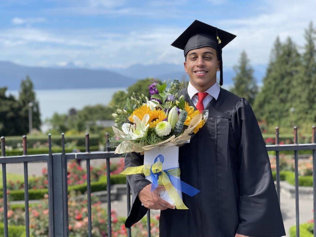 YOUNG GRADUATE WITH FLOWERS STANDING IN FRONT OF A FENCE